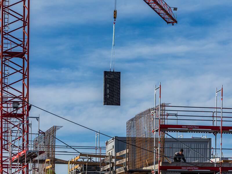 Ein Haus im Rohbau, das eingerüstet ist. Im linken Bildbereich befindet sich ein roten Kran,  welcher gerade Material an die Baustelle befördert.