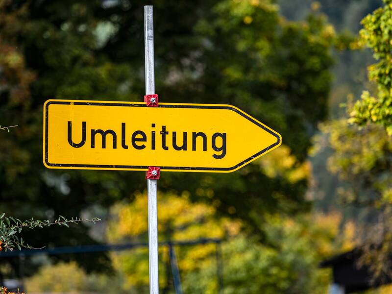 Ein gelbes Umleitungsschild, das in die rechte Richtung zeigt. Im Hintergrund sind herbstliche Bäume uns Sträucher.